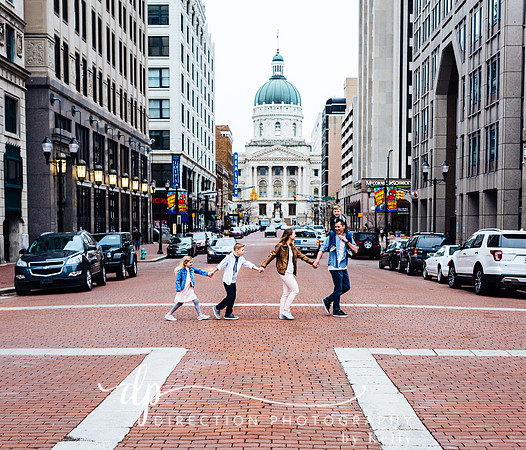 A family is crossing the street in downtown Indianapolis with the Indiana State Capital Building in the background.